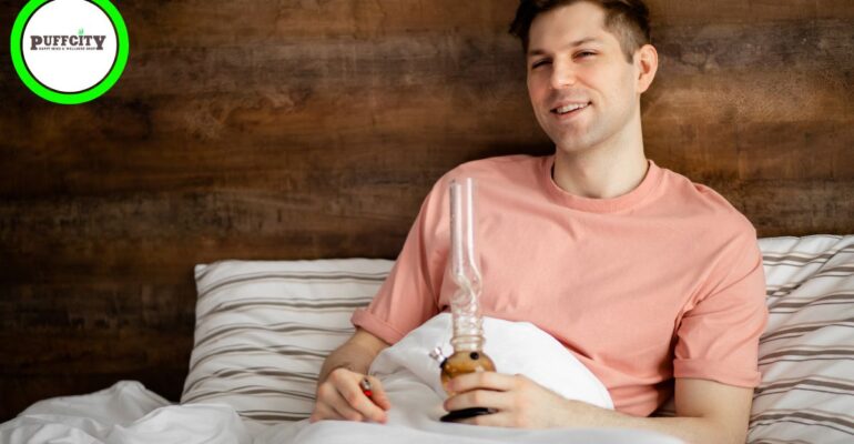 A man in a pink shirt with a bong in his hands is leaning on a bed.