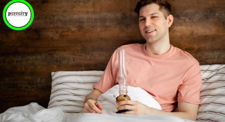 A man in a pink shirt with a bong in his hands is leaning on a bed.